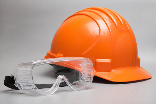 An orange hard hat and clear safety goggles are displayed against a plain gray background.