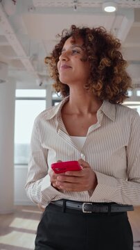 Vertical video of curly haired multiracial business woman in 30s walking with smartphone phone through office, texting message to client during short work break, medium shot of company employee.