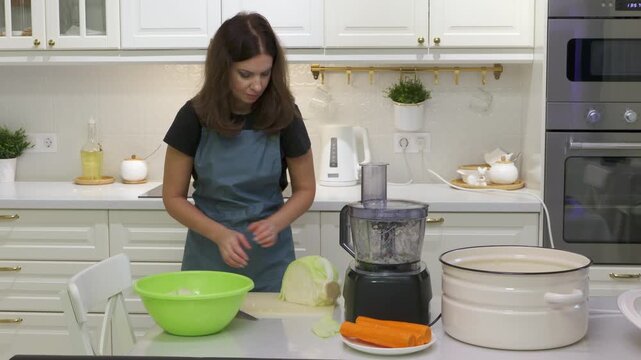 A woman in an apron cuts fresh cabbage at a table with carrots and a food processor nearby. She prepares healthy food calmly in a bright, modern white kitchen. She feels serene and focused.
