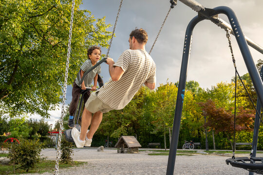 Caucasian father supporting his multiracial son on a playground swing during golden hour, representing racial diversity, attentive parenting, family bonding and outdoor childhood