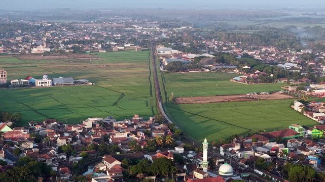 Sunlit aerial view of Kepanjen town with rice paddies, mosque minaret, and long road.