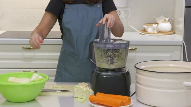 A woman wearing an apron uses a food processor to shred fresh cabbage. She prepares vegetables on a clean counter in a bright kitchen. Her calm focus creates a cozy and satisfying mood.