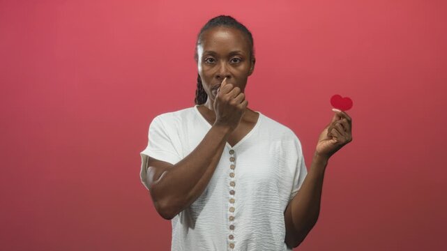 Young african american woman wearing white button shirt holds small red paper heart with right hand and finger to lips for silence in studio; quiet reflection.