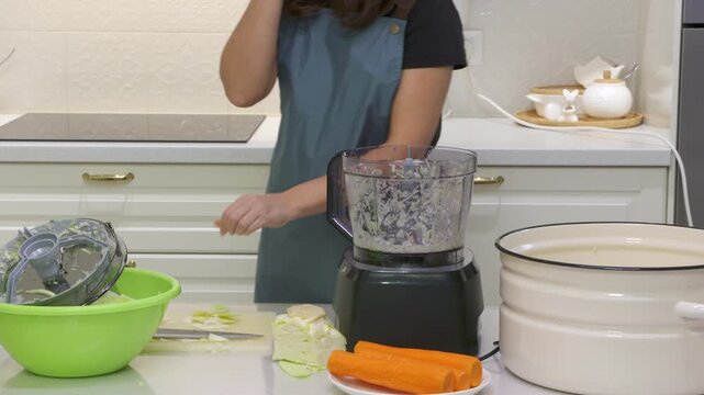 A woman in an apron opens the lid of a food processor. She prepares fresh cabbage and carrots at a clean kitchen counter. Her efficient moves bring a sense of calm and satisfaction.