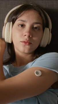 Vertical ugc shot of biracial teenage girl wearing headphones, showing glucose sensor on arm to camera. Footage fits youth diabetes awareness campaigns and healthcare ads