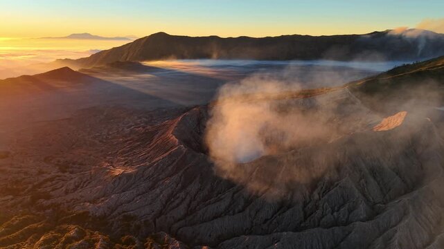 Aerial drone panorama of Mount Bromo at sunrise, mist filled crater glowing with golden light above clouds