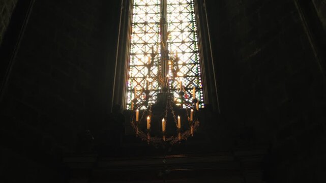 View of a tall stained glass window inside a historic Gothic cathedral. Sunlight shines through colorful glass, illuminating the sacred interior and highlighting medieval religious architecture.