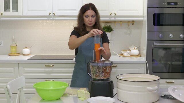 A woman in an apron pushes a carrot into a food processor. She prepares fresh food at a kitchen counter with focused care. Her smooth moves bring a calm and satisfying feeling.