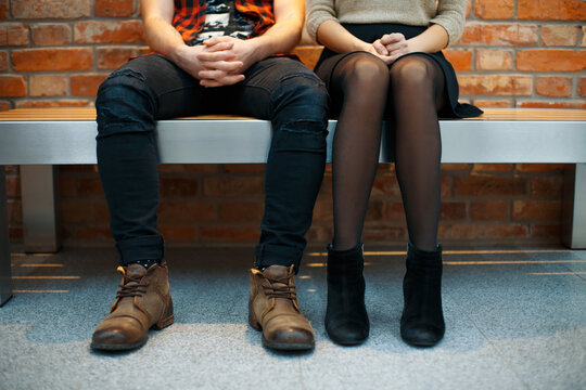 Cropped view of a young couple sitting side by side on a bench against a red brick wall. Man and woman in stylish clothes waiting together, concept of dating, relationship, and modern lifestyle.   