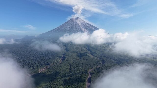Volcanic Semeru peak erupts, smoke rises into sky. Clouds drift around lush, green forest below