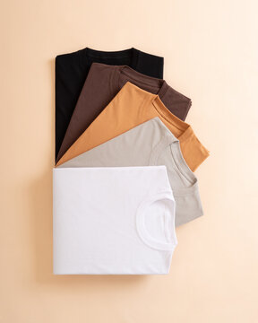 A collection of neatly folded t-shirts in various neutral earth tone colors arranged in a fan shape on a beige background