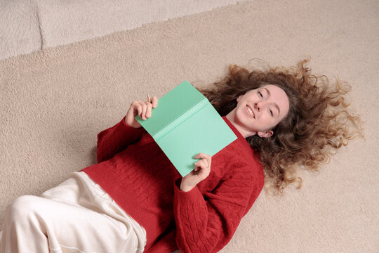 A smiling young woman with curly hair lying on the floor holding a green notebook for planning