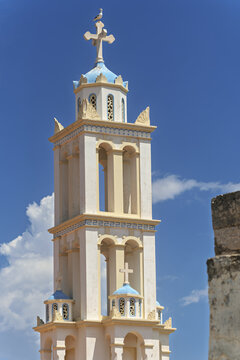 A blue dome and white cross of Church of Agios Nikolaos rise above the ornate Halki tower, with a lone gull perched on the ledge under clear island sky.