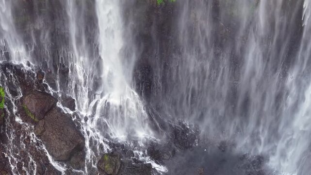 Powerful water crashes over jagged rocks below. Foam sprays everywhere as the torrent rushes forward at Tumpak Sewu