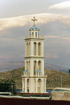 A blue dome and white cross of Church of Agios Nikolaos rise above the ornate Halki tower, with a lone gull perched on the ledge under clear island sky.
