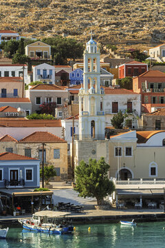 A blue dome and white cross of Church of Agios Nikolaos rise above the ornate Halki tower, with a lone gull perched on the ledge under clear island sky.
