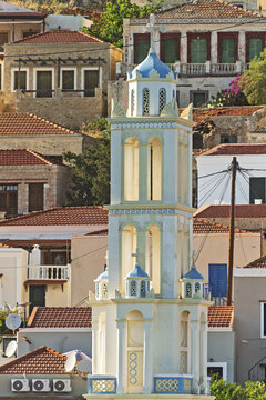 A blue dome and white cross of Church of Agios Nikolaos rise above the ornate Halki tower, with a lone gull perched on the ledge under clear island sky.