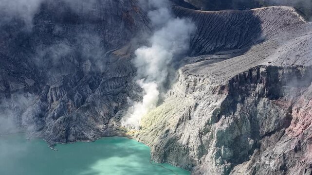 Volcanic smoke rises from a rugged crater. Turquoise waters surround the steep, rocky walls at Mount Ijen