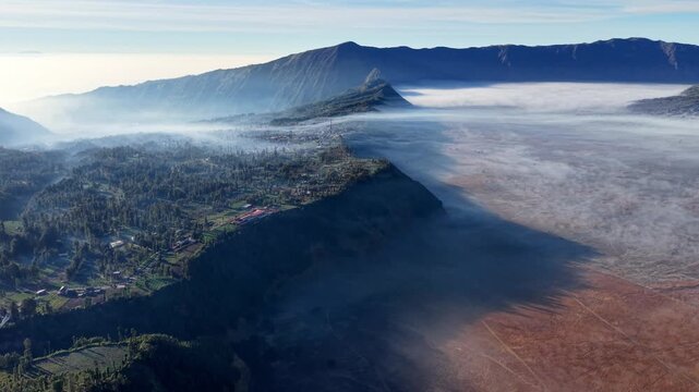 Misty peaks rise above lush green valleys. Dense fog curls around rugged, reddish slopes at Bromo