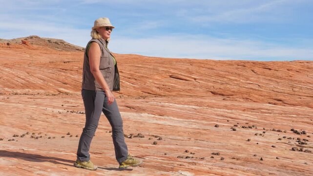 Side view of active mature woman in hiking gear walking on vibrant red rock formation under a clear blue sky at hot day in dried desert. Hiker enjoying an adventurous outdoor journey in national park