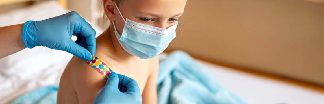 Doctor in blue gloves applying colorful adhesive bandage to shoulder of girl wearing protective face mask. Vaccination aftercare and pediatric treatment. Child health care and medical