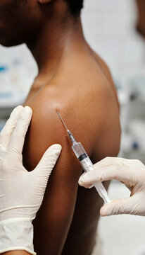 Doctor in sterile gloves injecting vaccine into bare shoulder of young man. Close-up of syringe needle entering upper arm. Immunization and disease prevention. Healthcare and medicine