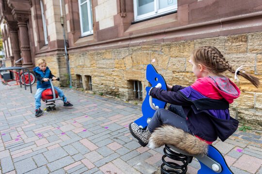 Two children enjoy spring riders on urban sidewalk beside historic stone building, sharing playful outdoor moment. Candid city scene captures childhood, friendship, recreation and active leisure