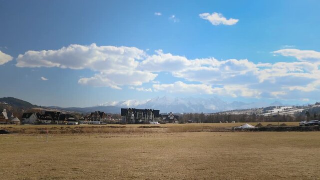 Wide-angle view of an empty countryside field with yellow grass, rolling hills, and bare trees beneath a bright blue sky filled with white fluffy clouds.