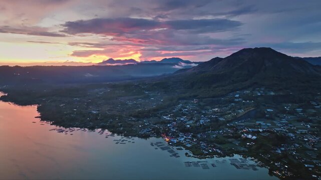 Amazing vibrant sunset over Mount Batur volcano, reflecting orange and purple hues across Lake Batur, scattered houses and fish farms dotting the Kintamani Bali landscape. Aerial view drone flight