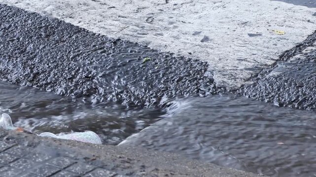 Rainwater entering storm drain during downpour on paved road. A small stream runs along the pavement toward a curb inlet. Urban water management, flooding risk and public infrastructure concept