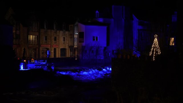 Nighttime static establishing shot of a picturesque riverside village decorated with glowing blue lights, holiday ornaments, and a golden Christmas tree. Historic stone, Pont Aven France