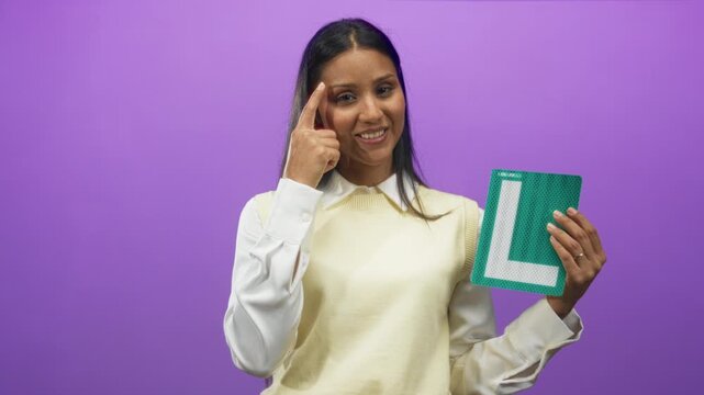 Woman points finger to temple while holding a green l learner plate, wearing white blouse and cream sweater in studio; confidence learning progress.