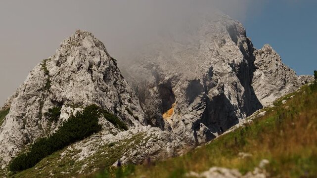 Low clouds drift around rugged mountain peaks in Slovenia's Kamnik Savinja Alps. Grass and shrubs cover the foreground below the steep rocky slopes.
