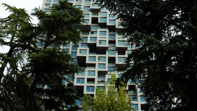 A modern tower with projecting square sections rises behind trees in Tirana, Albania. The close view highlights the geometric facade and rows of glass windows.