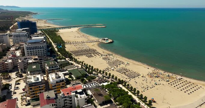 A drone view follows a busy beach in Durres, Albania, with rows of sun umbrellas and people beside the Adriatic Sea. Hotels line the developed waterfront.