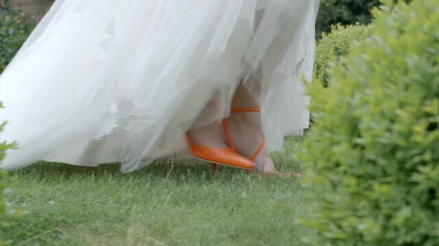 Close-up of bride walking on grass in white gown and bright orange high heels.