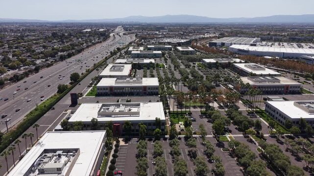 Drone tracking shot of a modern corporate office park and distribution center with a large parking lot next to a busy highway on a sunny day in Milpitas, California