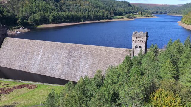 Aerial video the curved stone wall and Gothic towers of Derwent Dam surrounded by lush evergreen forests and rolling moorland hills of Derbyshire, UK. 