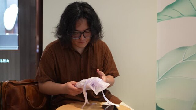 A man in glasses holds and inspects a translucent drawstring pouch while seated indoors at a cafe table.