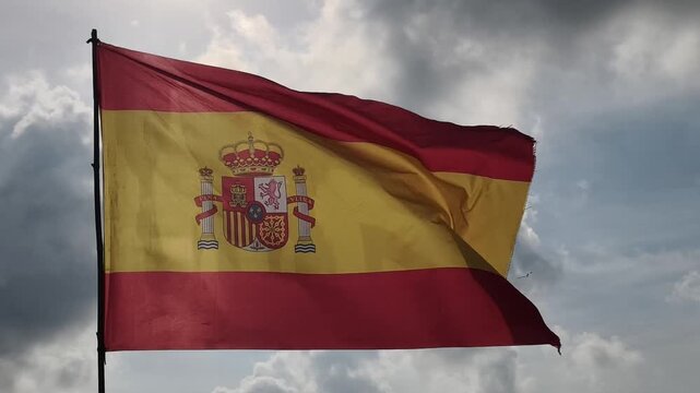 Spanish national flag waving dynamically against a cloudy sky. The bold red and yellow stripes feature Spain&rsquo;s detailed coat of arms with crowned shield and &ldquo;PLUS ULTRA&rdquo; columns. 
