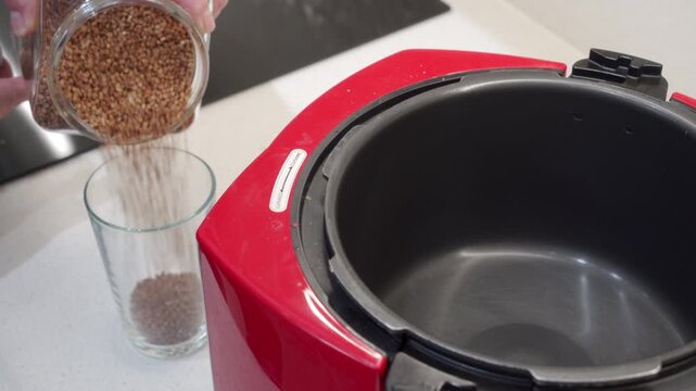 Hands pour raw buckwheat grains from a jar into a glass. A red multicooker stands ready on the clean kitchen counter. The neat meal preparation brings a cozy and satisfying feeling.