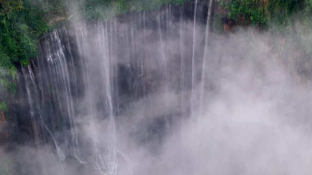 Mist rises from rushing water over dark cliff face. Greenery frames the cascade in misty haze at Tumpak Sewu