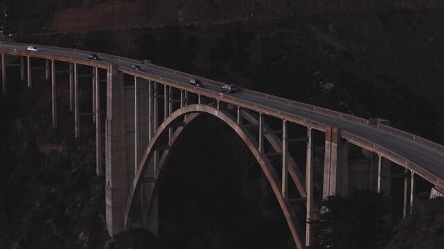 Aerial perspective of vehicles driving along the concrete arch structure of Bixby Creek Bridge on Highway 1 in Big Sur, California, surrounded by steep coastal cliffs during dusk.