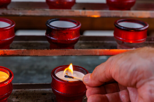 Close up of hand lighting a votive candle with a match in a red glass holder inside a church, religion and prayer concept