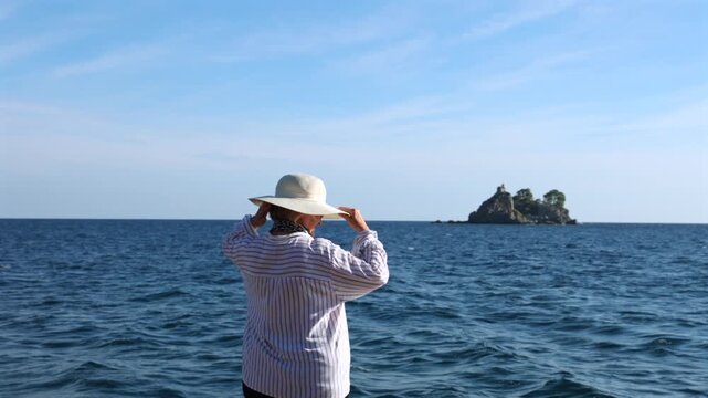 Senior Woman Adjusting Sun Hat and Looking at Sea During Retirement Travel. Senior woman in sun hat looking at the sea and distant island during a relaxing retirement vacation.