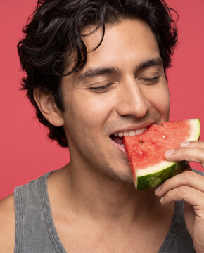 Refreshing man bites a slice of watermelon, with closed eyes against a bold color backdrop