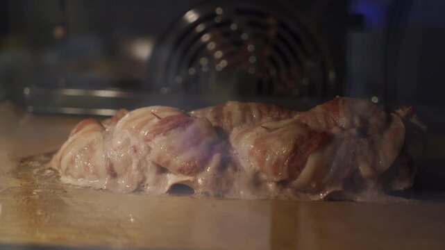 A bacon-wrapped meat roll bakes and sizzles on parchment paper inside a warm oven. Hot juices bubble around the savory roast. The appetizing meal brings a cozy, comforting, and satisfying feeling.