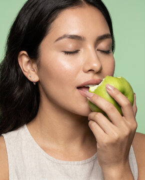 Crisp woman bites a fresh green apple, with closed eyes against a solid studio background