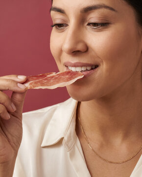 Refined woman bites a slice of cured ham, with elegance against a bold color backdrop