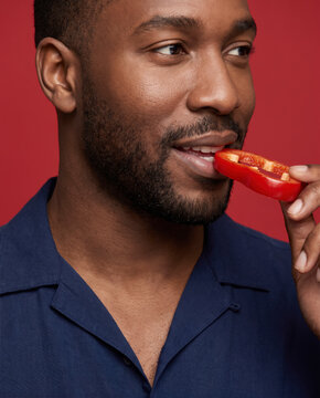Bold man bites a red bell pepper, with a smile against a solid studio background